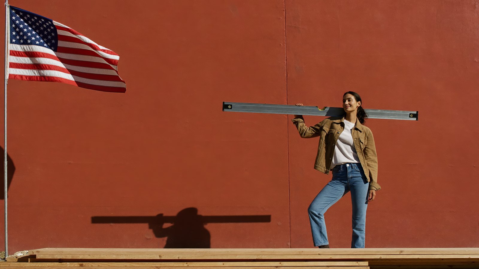Construction company owner carrying a long level on her shoulder, smiling toward the headline, a sunlit terracotta wall and an American flag beside her